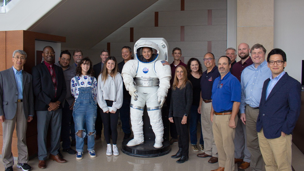 一群NASA代表、他们的家人和德州农工大学的教职员工站在一个真人大小的太空服模型周围，对着镜头微笑。