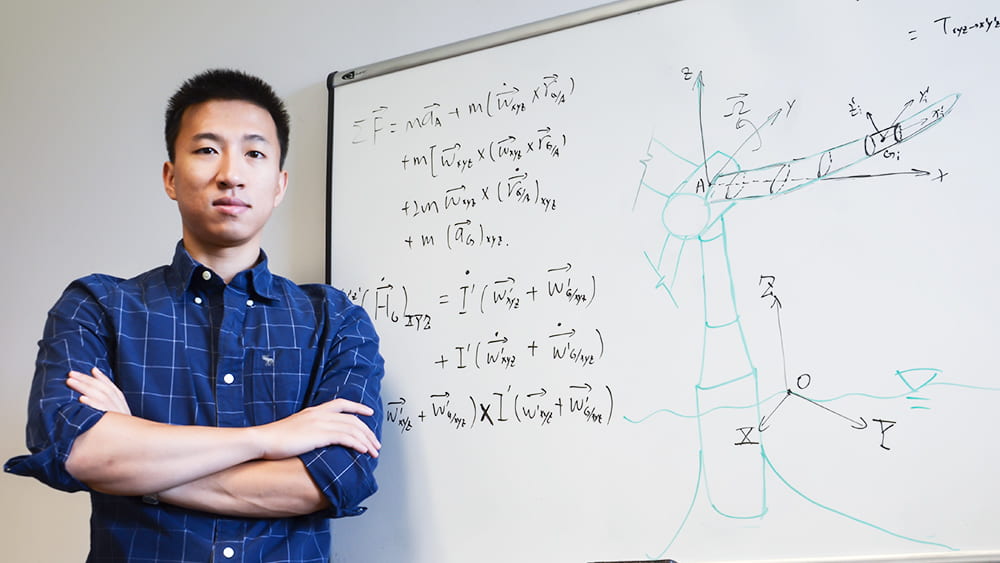 一名研究生站在一块白板前，上面写着方程式和图纸，代表他对海洋风力涡轮机的研究。