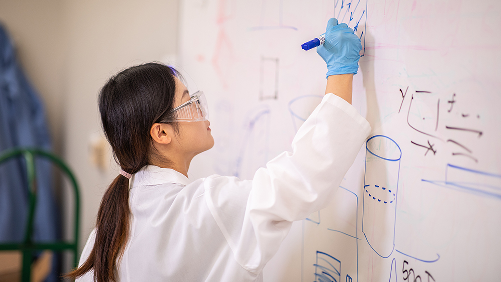 一名女学生穿着白色白大褂，戴着护目镜和蓝色手套，用蓝色记号笔在白板上写字。