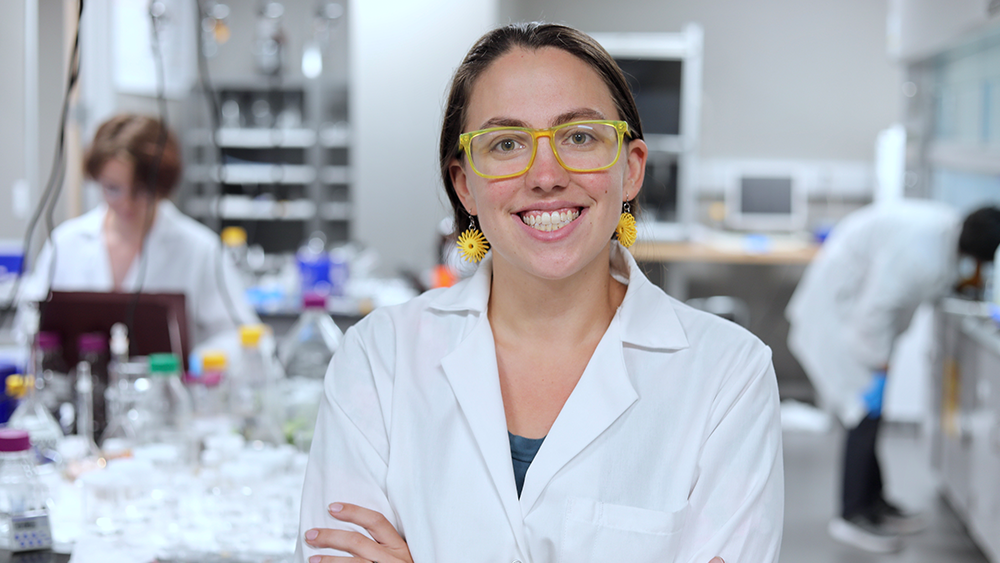 一名身穿白色实验服、戴着黄色眼镜的女子站在实验室里，双臂交叉微笑着