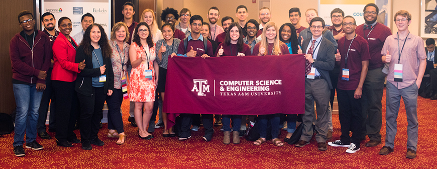 一群计算机科学和工程专业的学生和两位教授竖起大拇指。中间的学生举着印有系名和校徽的栗色横幅。他们在铺着红地毯和黄色圆点的会议室里。