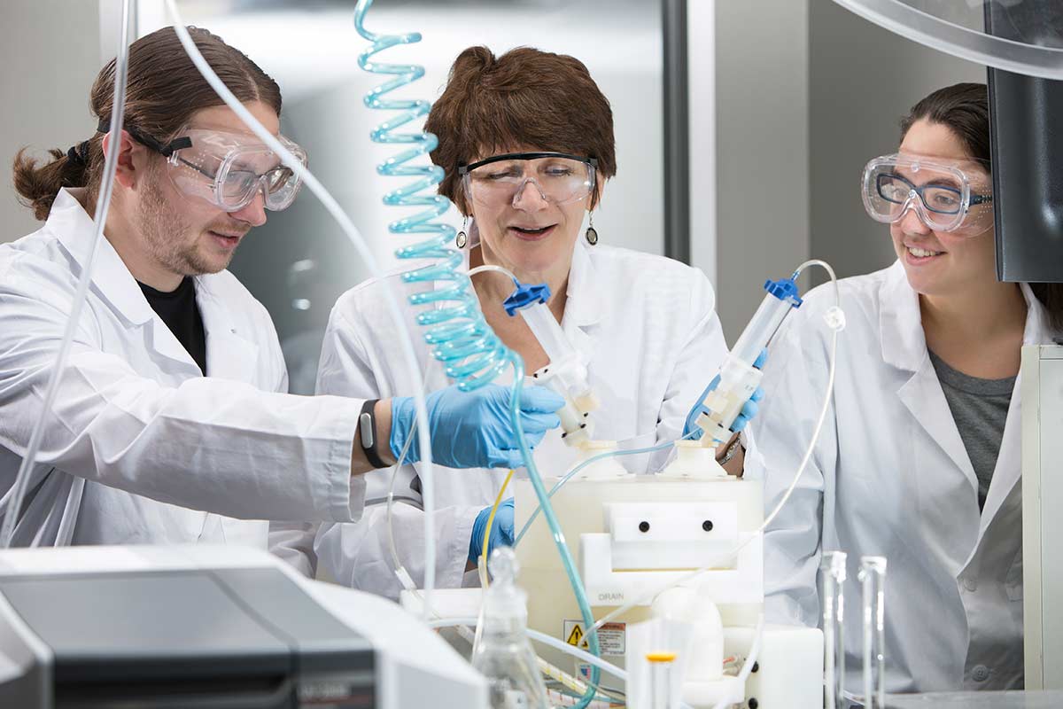 实验室里有两女一男。他们都穿着白色白大褂，戴着护目镜和蓝色塑料手套。