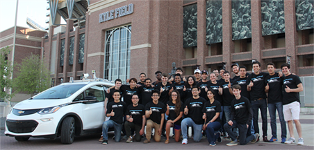 一大群身穿黑色t恤的学生(前排单膝跪地)站在凯尔球场砖墙外的白色汽车旁。