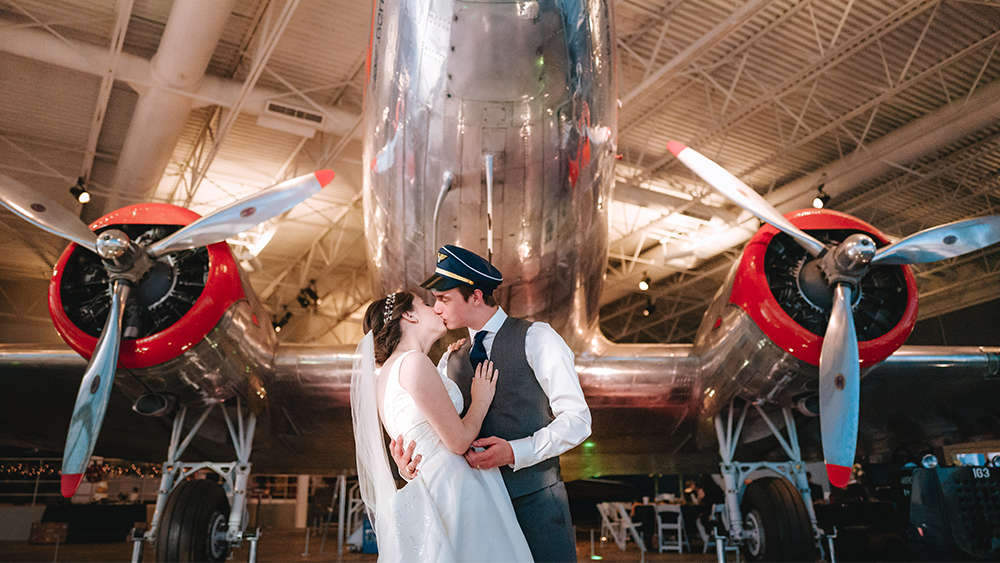 凯蒂和史蒂文以前是航空航天工程专业的学生，他们穿着新郎新娘的结婚礼服。凯蒂穿着一件带面纱的白色婚纱，史蒂文穿着一件深灰色背心，白色衣领衬衫，深蓝色领带，以及一顶深蓝色和金色的飞行员帽。史蒂文把凯蒂抱在怀里，凯蒂在一架经过修复的银红色DC-3飞机的鼻子下亲吻她。