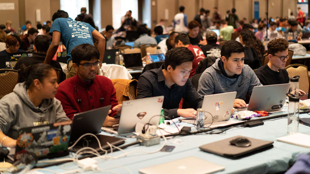 在TAMUhack活动中，四名学生坐在桌子上用笔记本电脑工作。