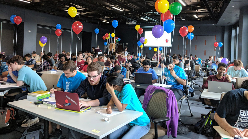 一个大房间里挤满了德州农工大学的学生，他们两三人一组坐在桌子旁，正在进行编程比赛。每张桌子上都有一台单独的、打开的笔记本电脑，学生们正在用它工作。一些椅子的靠背上绑着五颜六色的气球。