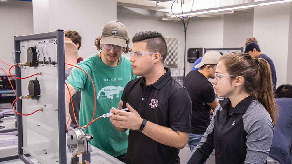 德克萨斯州农工大学的三名本科生和其他学生一起在实验室里用注射器和连接在板上的管子工作。