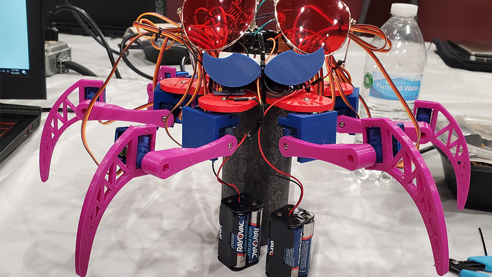 在一张桌子上，有六条腿的机器人Bitwise Boogie蓄着胡子，戴着太阳镜，下面用电线连接着两个电池组。