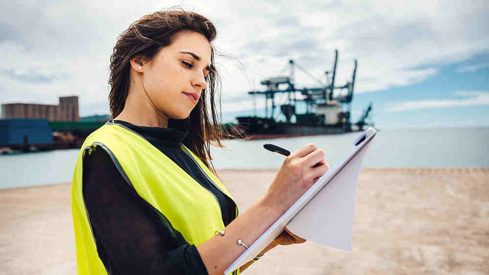 女工程师站在岸边，背景是一个石油平台，在图纸上做笔记。