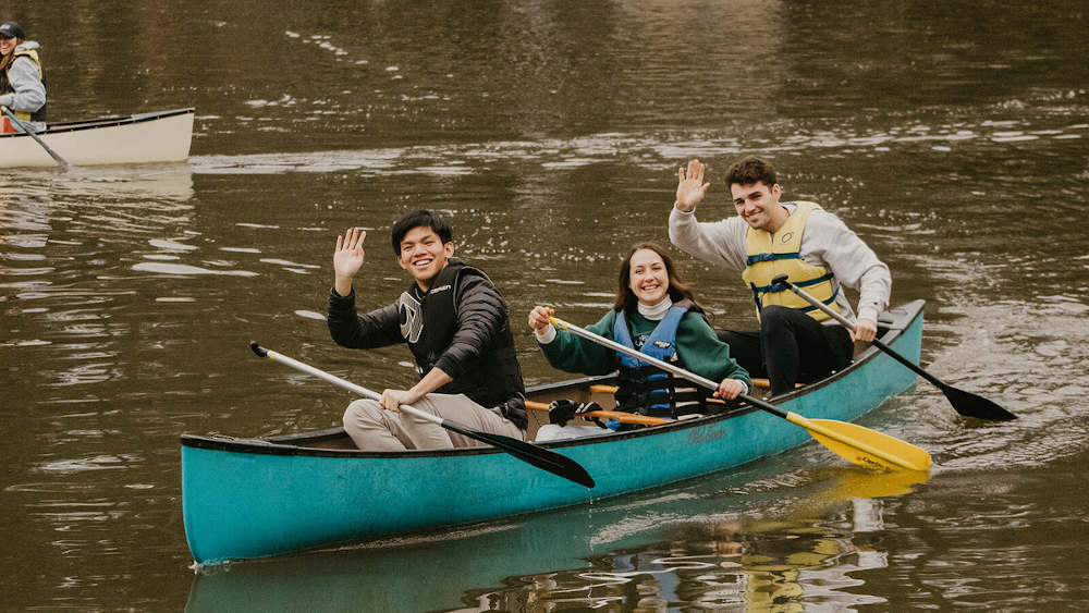 三个学生在湖面上的独木舟上向镜头挥手。